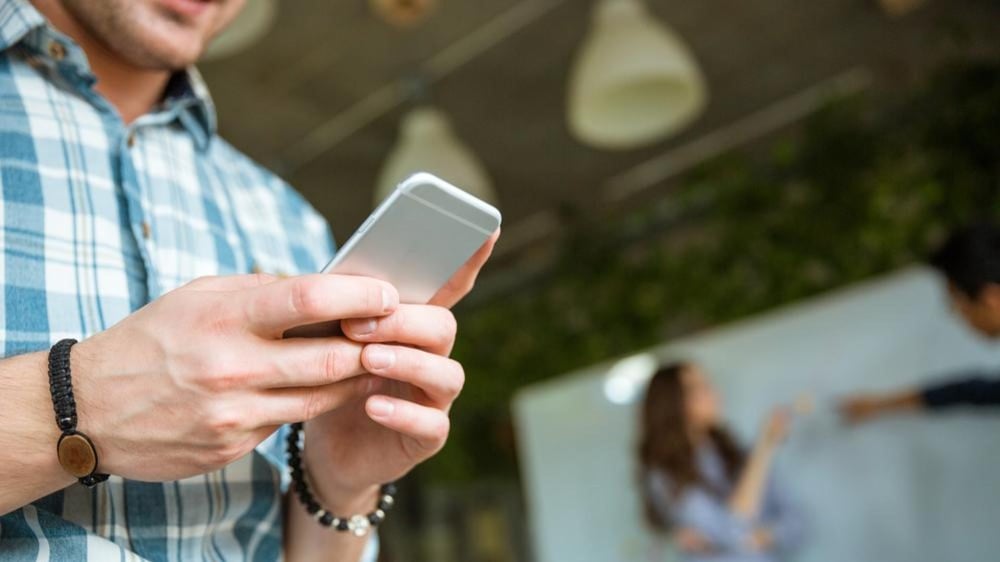 Closeup of hands of young man in checkered shirt using mobile phone while his partners arguing