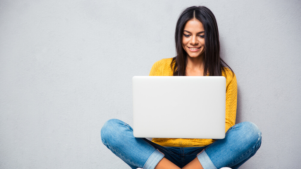 Happy young woman sitting on the floor with crossed legs and using laptop on gray background